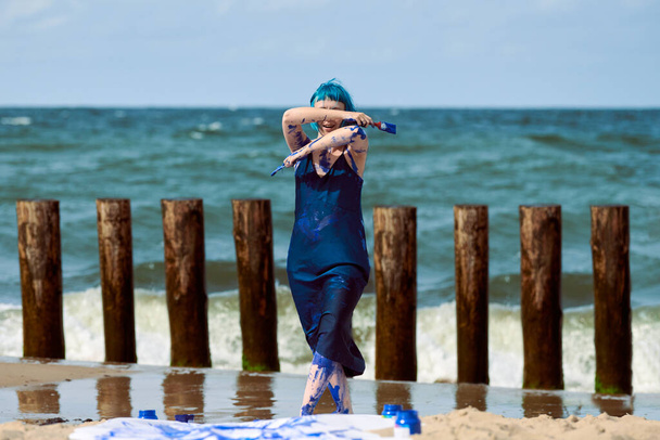 Artiste de performance femme aux cheveux bleus en robe bleu foncé enduite de peinture à la gouache bleue et dansant sur la plage. Body painting et concept d'art corporel, art de la performance expressif en plein air - Photo, image
