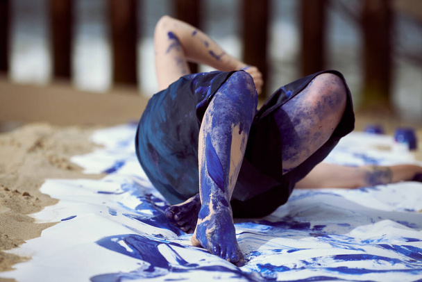 Female performance artist in dark blue dress smeared with blue gouache painting with wide strokes on large canvas lying on beach, close up. Expressive performance art, contemporary art, outside - Photo, Image