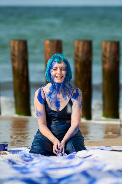 Artistic blue-haired woman performance artist in dark blue dress smeared with indigo gouache painting on her body with brushes on beach. Creative body painting, body art concept, outdoor performance - Photo, Image