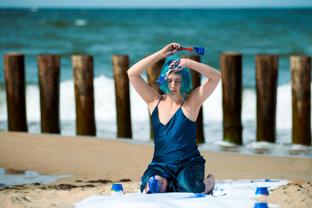 Artiste de performance femme aux cheveux bleus en robe bleu foncé enduite de peinture à la gouache bleue alors qu'elle était assise sur la plage. Body painting et concept d'art corporel, art de la performance expressif en plein air - Photo, image