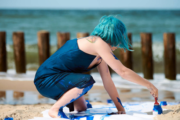 Mavi saçlı, koyu mavi elbiseli, kumsaldaki büyük tuvale geniş dokunuşlarla mavi guaj boyalı bir kadın performans sanatçısı. Etkileyici performans sanatı, modern açık hava sanatı. - Fotoğraf, Görsel