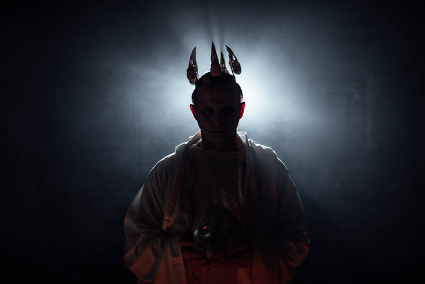 a man in the makeup of a dead man and a crown on his head. a gloomy photo shoot with special effects of smoke and light. cosplay a demon for Halloween. the silhouette of a man in the dark - Photo, Image