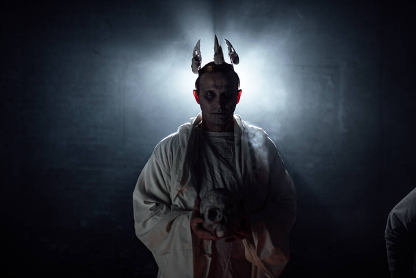 死んだ男の化粧をした男と頭に王冠。煙と光の特殊効果で暗い写真撮影。ハロウィンの悪魔コスプレ。暗闇の中の男のシルエットは - 写真・画像