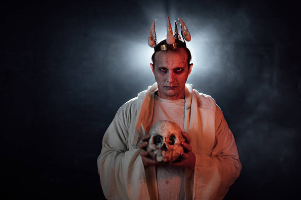 死んだ男の化粧をした男と頭に王冠。煙と光の特殊効果で暗い写真撮影。ハロウィンの悪魔コスプレ。暗闇の中の男のシルエットは - 写真・画像