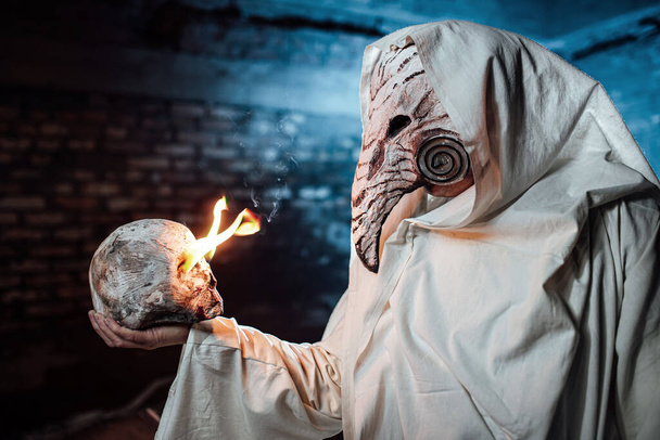 Een man verkleed als een pest dokter houdt een menselijke schedel in zijn hand. gekostumeerd feest voor Halloween. cosplay van de ruiter van de Apocalyps. rekwisieten brandende schedel - Foto, afbeelding