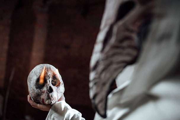 Ein als Pestarzt verkleideter Mann hält einen menschlichen Schädel in der Hand. Kostümparty zu Halloween. Cosplay des Reiters der Apokalypse. Requisiten verbrennen Schädel - Foto, Bild