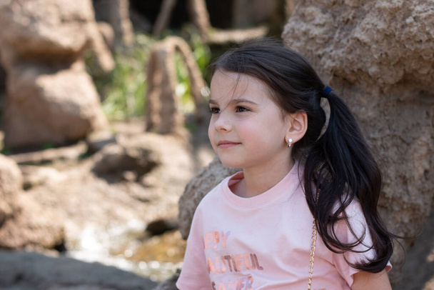Malá krásná bílá dívka s tmavými dlouhými vlasy na pozadí zahrady z kamení - Fotografie, Obrázek