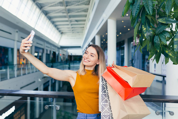 young happy after shopping woman talking on cell in mall. Satisfied joyful female shopaholic with gift colored paper bags in hands. Mobile phone conversation. Smiling joy Stand ang sincerely tells - Foto, afbeelding