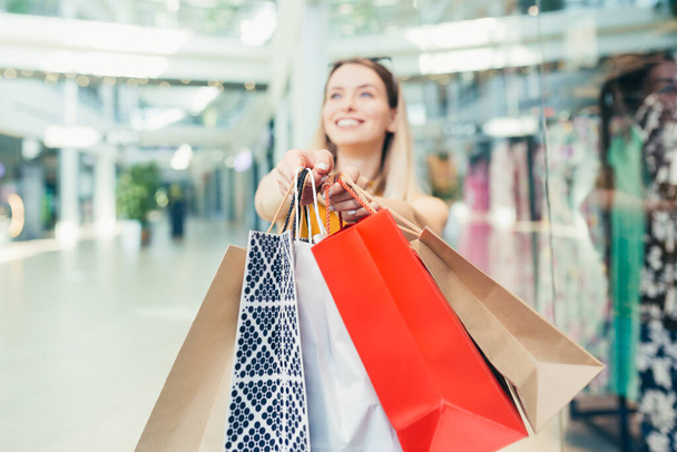 cheerful young shopaholic woman holding paper bags with purchases and smiling looking at camera dancing on shopping center. Happy lady having fun on black friday joyfully jumps for joy in the mall - Φωτογραφία, εικόνα