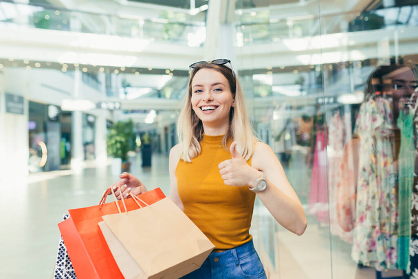 cheerful young shopaholic woman holding paper bags with purchases and smiling looking at camera dancing on shopping center. Happy lady having fun on black friday joyfully jumps for joy in the mall - Foto, afbeelding