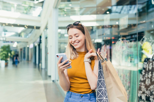 Young woman consumer in the mall browses chat and uses using a smartphone. female standing with a mobile phone in her hands in shopping center. indoor. happy shopper girl with gift bags make purchases - Zdjęcie, obraz