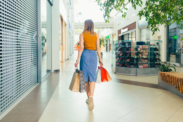 Rear Back view of a young woman with colored gift bags walking down the hallway of a mall or store. Satisfied happy blonde female shopper while shopping indoors. Go and look around the camera - 写真・画像
