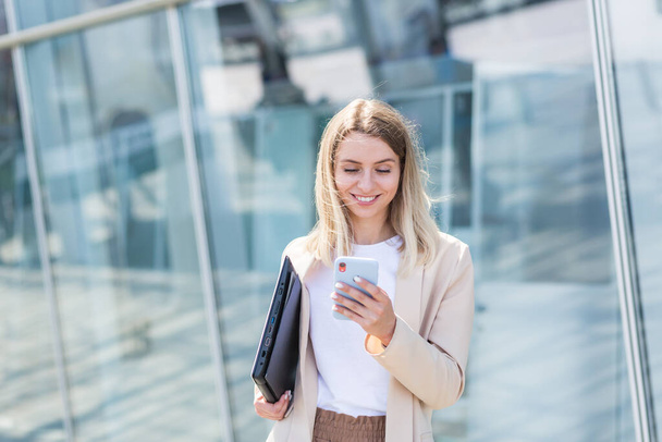 young business woman goes and uses mobile phone in her hands a urban background a modern office building. Attractive businesswoman or student Female walks the city street outdoors using a smartphone - Foto, Imagen