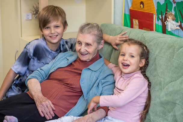 Szczęśliwa rodzina - babcia z wnukami, dziewczynka i chłopiec przytulają babcię, wszyscy razem siedzą na kanapie w pokoju, więzi rodzinne, dzieciństwo, młodość i starość. - Zdjęcie, obraz