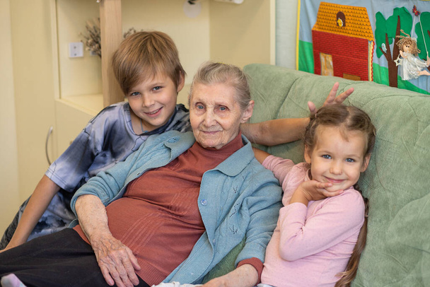 Een gelukkig gezin - een grootmoeder met kleinkinderen, een meisje en een jongen knuffelen hun grootmoeder, allemaal samen zitten mensen op de bank in de kamer, familiebanden, jeugd, jeugd en ouderdom. - Foto, afbeelding