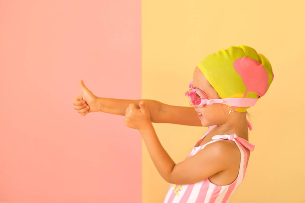 Chica positiva alegre en un traje de baño a rayas de moda, gafas de natación y una gorra de la piscina que muestra gesto de éxito, pulgar hacia arriba con la mano, diciendo bien hecho. Fondo de estudio rosa-amarillo. Super cool natación - Foto, Imagen
