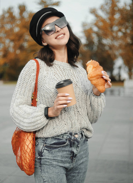 молодая женщина в оранжевом свитере с круассаном и чашкой кофе  - Фото, изображение