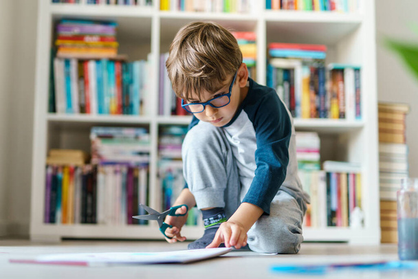 Vista frontal de un niño caucásico de cuatro años jugando n el suelo en casa - niño pequeño usando tijeras para cortar la educación de papel desarrollando habilidades artísticas creciendo concepto - Foto, imagen