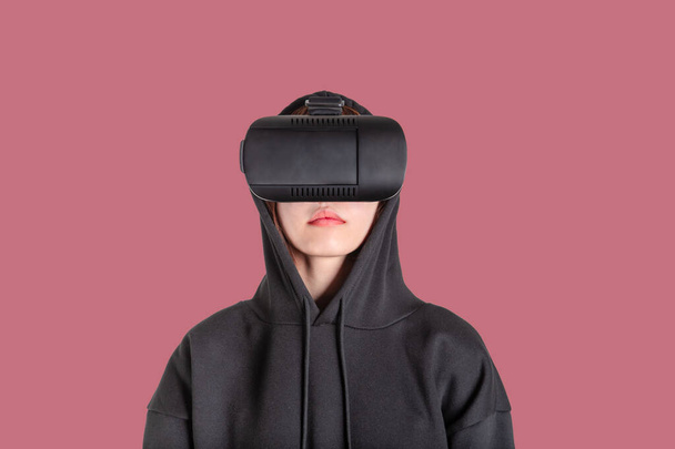 Junge Asiatin in schwarzem Kapuzenpulli mit VR-Gerät oder Virtual-Reality-Brille vor rosa Hintergrund. - Foto, Bild