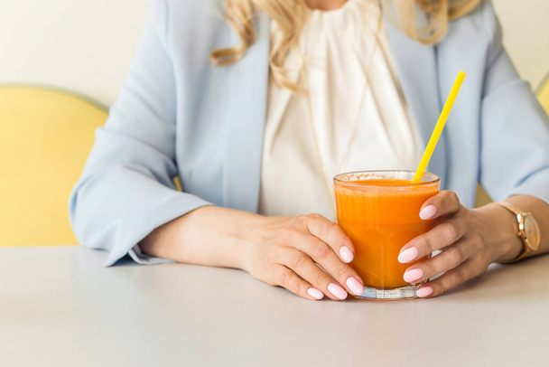 Um copo de suco de cenoura recentemente espremido nas mãos de uma jovem mulher em uma jaqueta azul e blusa branca. Foto - Foto, Imagem