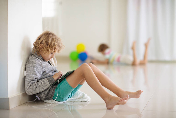 Kid with tablet computer. Child playing game on tile floor in white room. Kids no communicate at birthday party, gaming and screen time addiction. Little boy watching movie, chatting with friends - 写真・画像