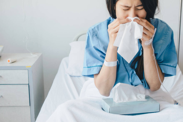 Portrait of young woman wearing patient cloths sneezing in tissue at hospital. Conceptual of sickness woman feeling when admitted in the hospital. - Photo, Image