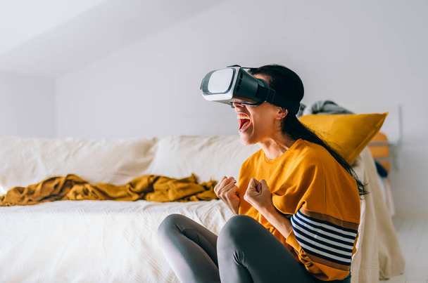 Podekscytowana kobieta odzieżowa w okularach VR wirtualnej rzeczywistości w salonie. Przyszły trend przy użyciu wirtualnej rzeczywistości gogle urządzenia do gier wideo i rozrywki domowej. Futurystyczne doświadczenie technologiczne. - Zdjęcie, obraz