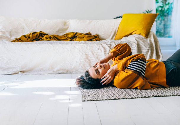 Woman in casual clothing with headphones lying down the floor in her living room and enjoys listening the music. Leisure time of single woman relaxing in isolation streaming songs from player.  - 写真・画像