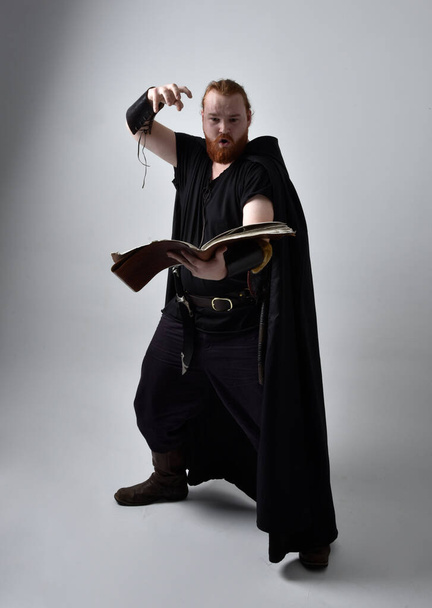  Porträt eines rothaarigen Mannes in einem mittelalterlichen Wikingerkostüm mit Mantel. Stehende Pose mit einem magischen Buch, mit gestischen Handbewegungen, als würde sie verzaubern, isoliert vor Studiohintergrund. - Foto, Bild
