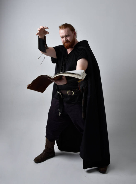  portrait d'un homme aux cheveux roux portant un costume fantaisie inspiré du viking médiéval avec manteau. pose debout tenant un livre magique, avec des mouvements gestuels de la main comme un sort, isolé sur fond de studio. - Photo, image