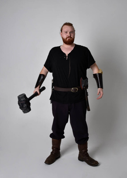  portrét rudovlasého muže ve středověkém vikingském kostýmu inspirovaném fantazií. Stojící póza s gesturálními pózami držící kladivo jako zbraň, izolovaná na pozadí studia. - Fotografie, Obrázek