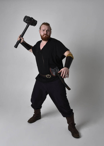  Portret rudego mężczyzny w średniowiecznym kostiumie wikinga inspirowanym fantazją. Pozycja stojąca z gestykulacyjnymi pozycjami trzymającymi młotek jako broń, odizolowana na tle studia. - Zdjęcie, obraz