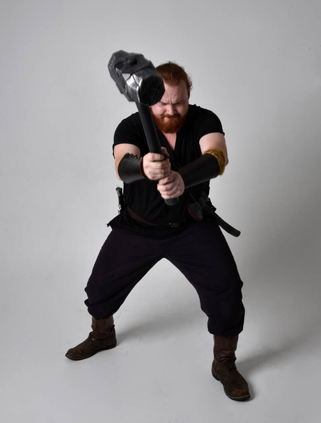 portrait of red haired man wearing medieval viking inspired fantasy costume. Standing pose with gestural poses holding a hammer as weapon, isolated  against studio background. - Foto, Imagem