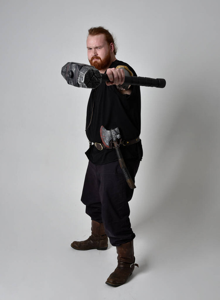  portrait of red haired man wearing medieval viking inspired fantasy costume. Standing pose with gestural poses holding a hammer as weapon, isolated  against studio background. - Foto, imagen