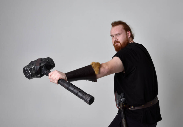  portrait of red haired man wearing medieval viking inspired fantasy costume. Standing pose with gestural poses holding a hammer as weapon, isolated  against studio background. - Fotoğraf, Görsel