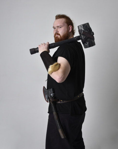  portrait of red haired man wearing medieval viking inspired fantasy costume. Standing pose with gestural poses holding a hammer as weapon, isolated  against studio background. - Photo, Image