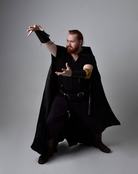  Portret rudego mężczyzny w średniowiecznym kostiumie inspirowanym wikingiem z ciemnym płaszczem. Postawa stojąca z gestykulacyjną ręką, jakby rzucała magiczne zaklęcie, odizolowana na tle studia. - Zdjęcie, obraz
