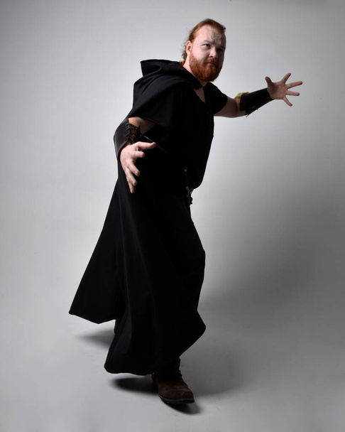  Portret rudego mężczyzny w średniowiecznym kostiumie inspirowanym wikingiem z ciemnym płaszczem. Postawa stojąca z gestykulacyjną ręką, jakby rzucała magiczne zaklęcie, odizolowana na tle studia. - Zdjęcie, obraz