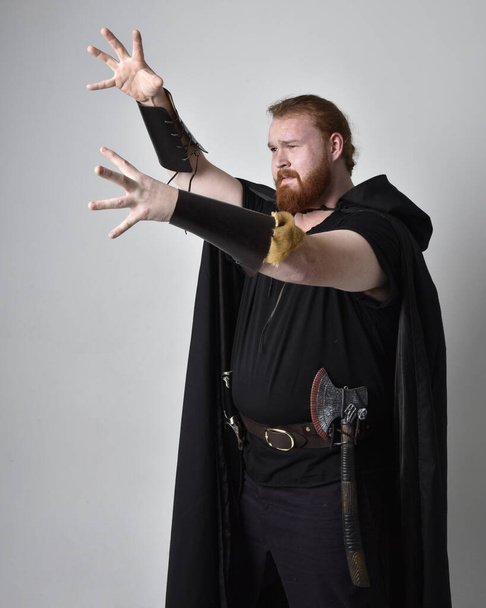  portrait of red haired man wearing medieval viking inspired fantasy costume with dark flowing cloak. Standing pose with gestural  hand poses, as if casting a magical spell, isolated  against studio background. - Foto, Imagen