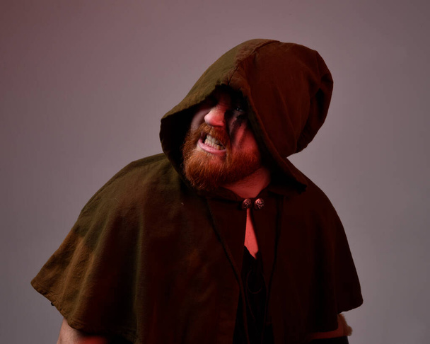  πορτρέτο του κοκκινομάλλη φορώντας μεσαιωνική Βίκινγκ εμπνευσμένο κοστούμι μανδύα φαντασίας με σκούρες σκιές και το πρόσωπο χρώμα. με φόντο σκοτεινό στούντιο με φωτισμό σιλουέτας. - Φωτογραφία, εικόνα