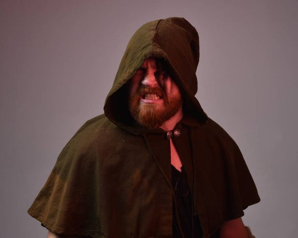  portret van roodharige man in middeleeuwse viking geïnspireerd fantasie mantel kostuum met donkere schaduwen en gezicht verf. tegen een donkere studio achtergrond met silhouet verlichting. - Foto, afbeelding