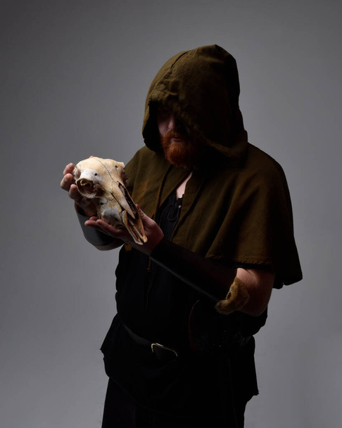  portrait of red haired man wearing medieval viking inspired fantasy costume with hooded cloak and face paint, holding a  sheep skull in pagan ritual,  studio background with silhouette rim lighting. - Foto, Imagen
