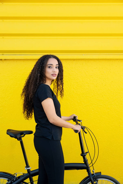 Stílusos fiatal fekete nő hosszú hajjal, aki a hajtogatott kerékpárját cipeli a sárga fal előtt - Fotó, kép