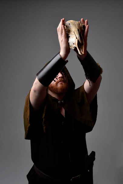  портрет рыжеволосого человека в средневековом костюме викинга с капюшоном и краской на лице, держащего череп овцы в языческом ритуале, студийный фон с силуэт-краем освещения. - Фото, изображение