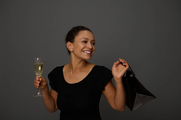 Krásná žena nakupující oblečený v černých šatech se usmívá při pohledu na kopírování prostor na šedém pozadí se sklenkou sarkling vína a nakupování černý balíček. Koncepce oslav Černý pátek - Fotografie, Obrázek