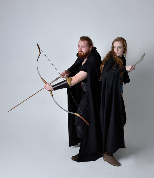 Teljes hosszúságú portré vörös hajú pár, férfi és nő visel középkori viking ihlette fantázia jelmezek, álló harci póz tartó íj és nyíl és hosszú kard fegyverek, elszigetelt fehér stúdió háttér. - Fotó, kép