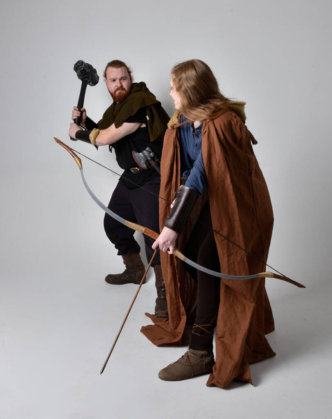 Full length  portrait of red haired  couple, man and woman wearing medieval viking inspired fantasy costumes, standing fighting pose holding  archery bow and arrow and long sword weapons, isolated on white  studio background. - Photo, Image
