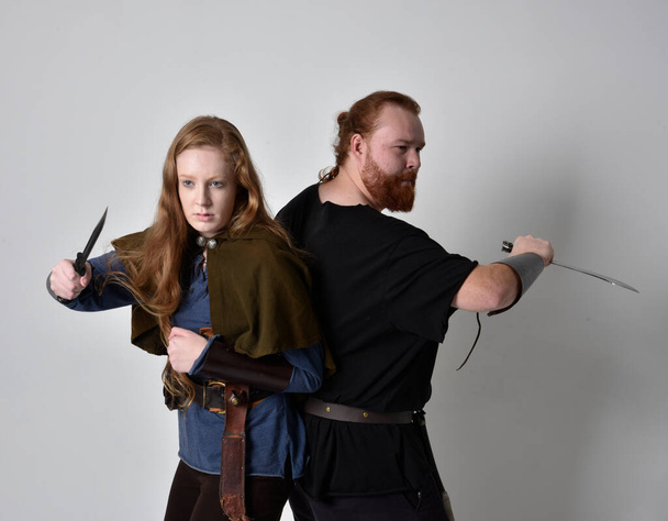 Полноразмерный портрет рыжеволосой пары, мужчины и женщины, одетые в костюмы средневековых викингов, вдохновленные фантазией, стоящие в боевой позе с оружием в руках, изолированные на белом фоне студии. - Фото, изображение