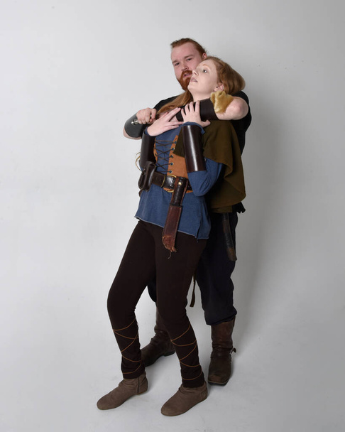 Πλήρες πορτραίτο του ζευγαριού με τα κόκκινα μαλλιά, άντρας και γυναίκα που φορούν μεσαιωνικά viking εμπνευσμένα κοστούμια φαντασίας, στέκονται πολεμώντας θέτουν κρατώντας όπλα σπαθί, απομονώνονται σε λευκό φόντο στούντιο. - Φωτογραφία, εικόνα