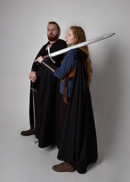 Ganztägiges Porträt eines rothaarigen Paares, Mann und Frau in mittelalterlichen Wikinger inspirierten Fantasiekostümen, stehend kämpfend mit Schwertwaffen, isoliert auf weißem Studiohintergrund. - Foto, Bild